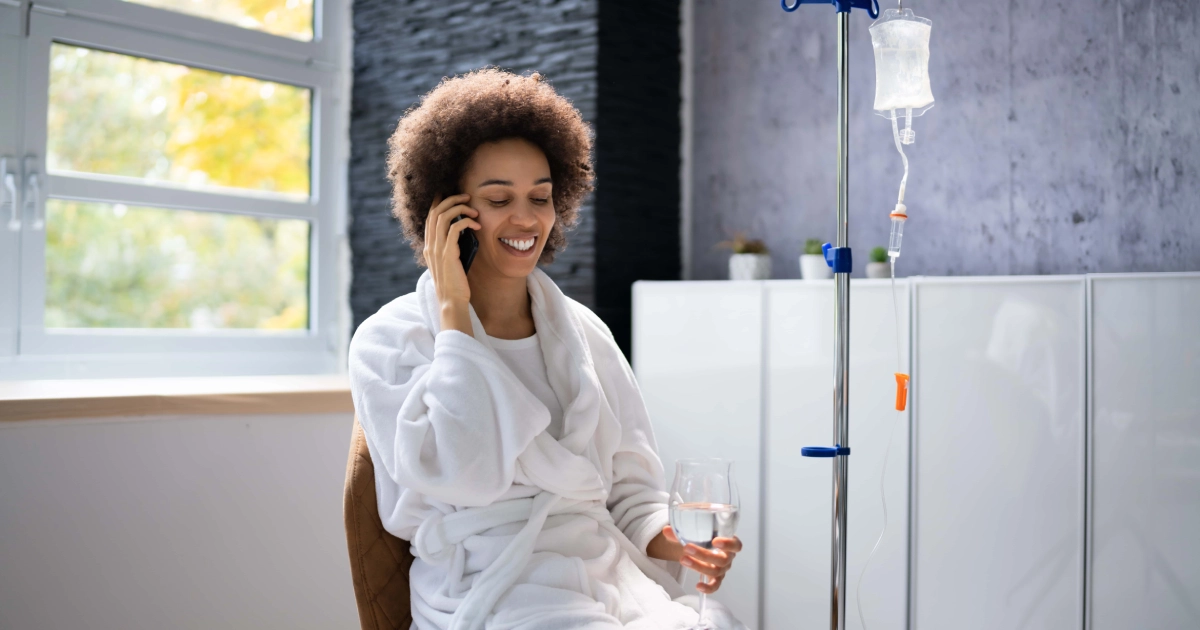 A woman in a white bathrobe sits next to an IV drip, smiling while talking on a smartphone and holding a glass of water during IV Hydration Therapy in Salado, TX.