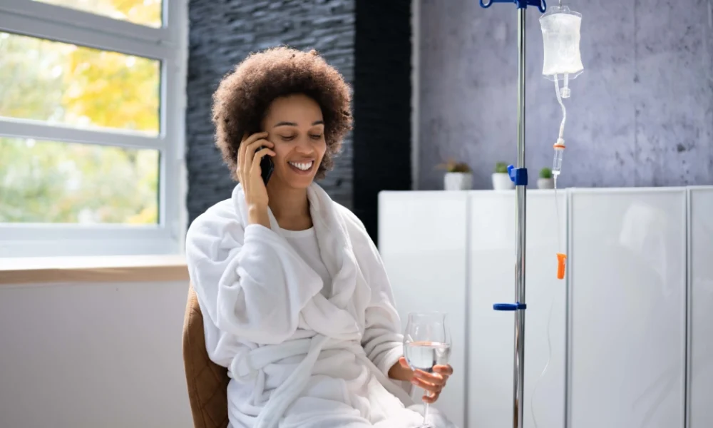 A woman in a white bathrobe sits next to an IV drip, smiling while talking on a smartphone and holding a glass of water during IV Hydration Therapy in Salado, TX.